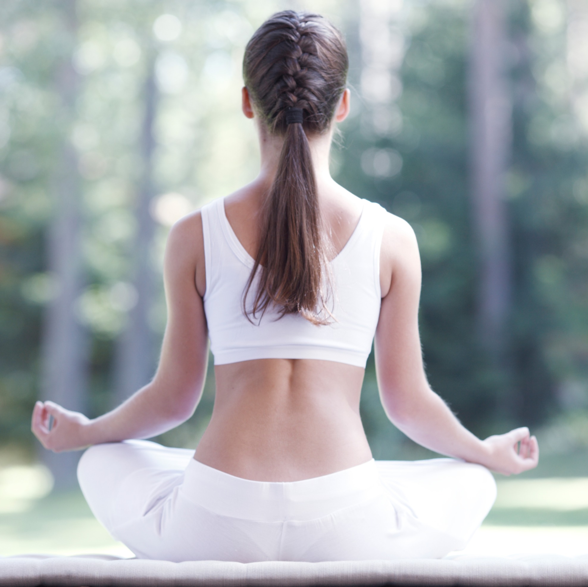 back of woman with partially braided brown hair and white exercise clothing while meditating cross legged in forest setting